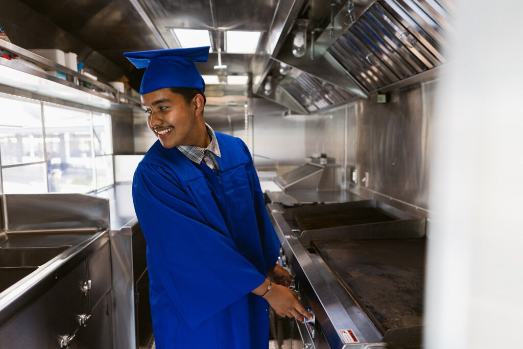 Giovani, a Polytechnic High School senior, wearing his graduation gown while standing in his food truck kitchen